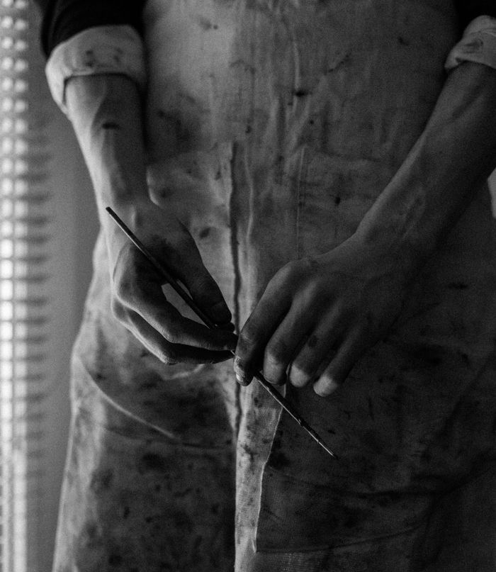 Black and white close-up of artist's hands holding a paintbrush in a studio setting.