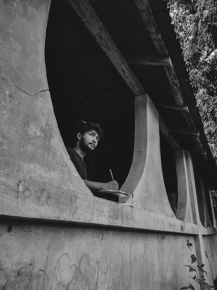 An Asian man thoughtfully writes in a notebook by a concrete window, grayscale image.
